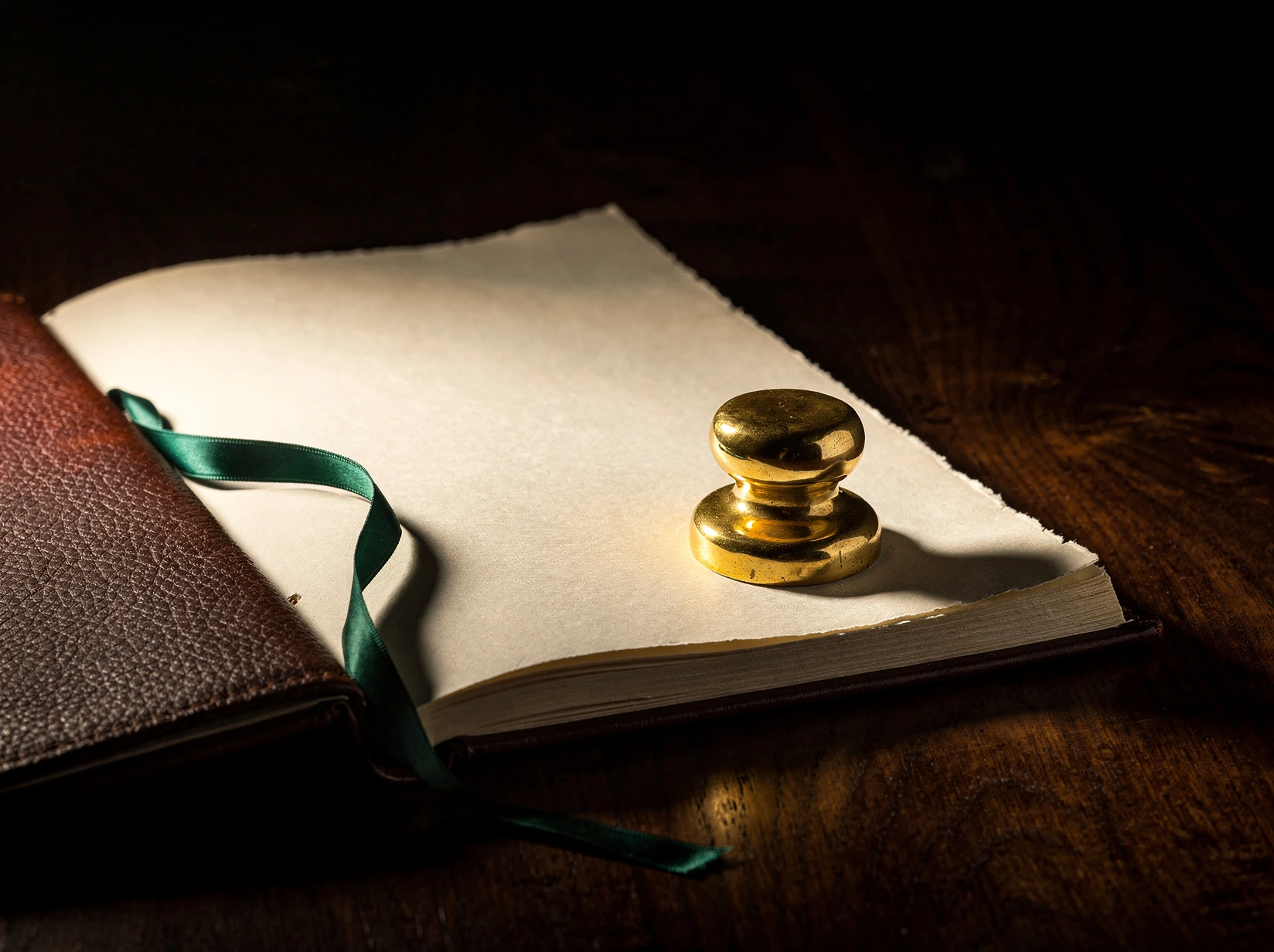 An open leather-bound ledger with cream blank pages, a polished brass weight resting on the right page, a deep forest green silk ribbon trailing across, on a black walnut tabletop, lit by a single warm tungsten lamp.